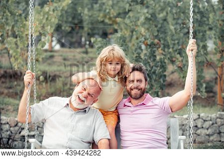 Boy Son With Father And Grandfather Swinging Together In Park Outdoors. Three Different Generations 