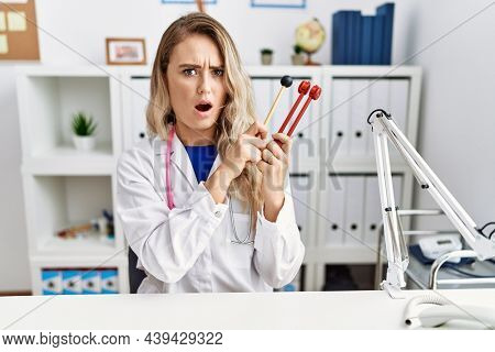Young beautiful doctor woman holding diapason instrument in shock face, looking skeptical and sarcastic, surprised with open mouth 
