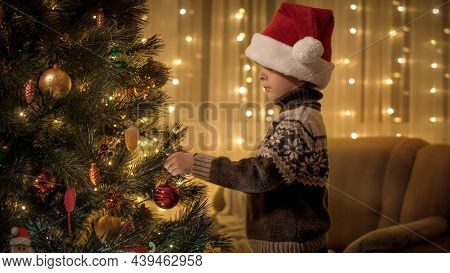 Portrait Of Little Boy Hanging Red Bable On Glowing Colorful Christmas Tree In Living Room. Pure Emo
