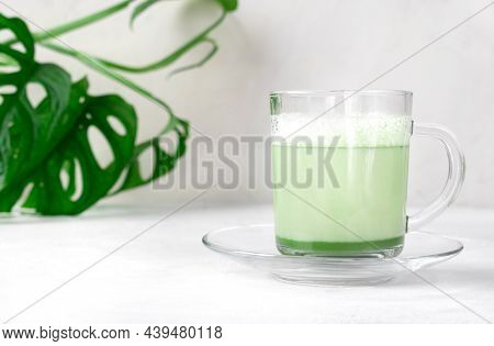 Matcha Latte With Foam On Plant-based Milk In Glass Cup On The White Desk With Monstera Houseplant. 