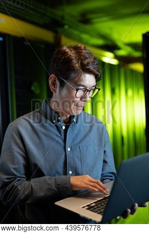 Asian male engineer using laptop in computer server room. database server management and maintenance concept