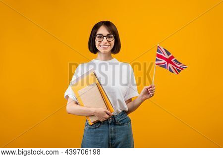 Travel And Study, Learn English Language. Zoomer Smiling Woman With Notebooks And Small Flag, Orange