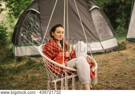 Beautiful Girl Wrapped In Red Plaid Drinking Tea In A Cozy Hanging Chair Outdoors. Girl Holding Mug 