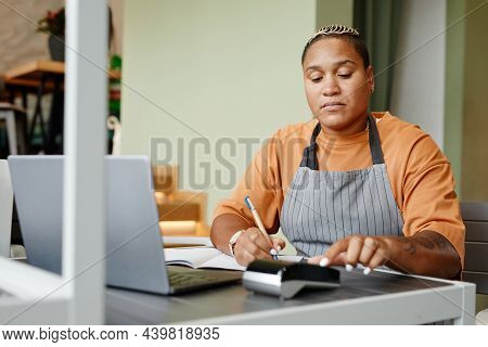 Young Woman Wearing Apron Sitting At Table In Front Of Laptop In Modern Cafe Taking Online Orders Fo