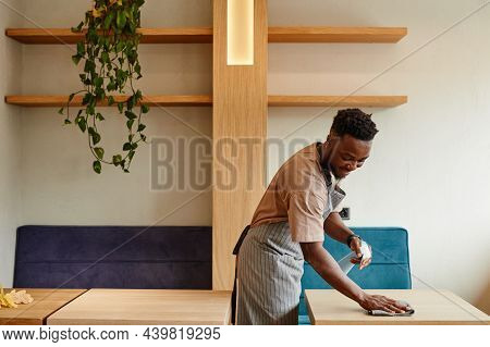 Medium Long Shot Of Young African American Man Wearing Apron Working In Small Cafe Preparing Tables 