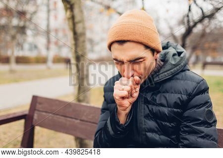 Young Sick Man Coughing Sitting On Bench In Fall Park Covering Mouth. Coronavirus Covid-19 Pandemic 