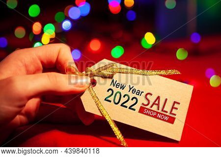A Womans Hand Holds A Label With Text - New Year Sale 2022, Shop Now. On A Red Background, With A Mu