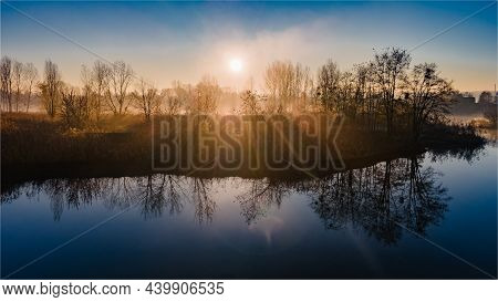 The Rays Of The Morning Sun Shining Through The Autumn Trees And Reflected In The Water Of The Misty