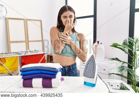 Young hispanic woman ironing clothes at laundry room smelling something stinky and disgusting, intolerable smell, holding breath with fingers on nose. bad smell 