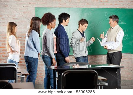 Professor gesturing thumbsup to successful male student with classmates standing in a row at classroom