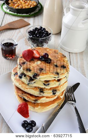 High angle shot of a stack of fresh homemade blueberry pancakes. The plate is on a rustic farmhouse style kitchen table. Other items include a syrup pitcher, knife, fork, skillet with fried potatoes.