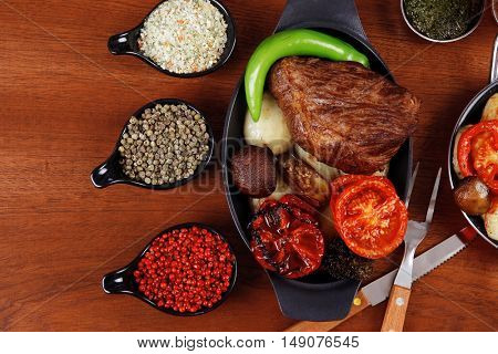 fresh roast beef meat rib eye steak with tomatoes,potatoes, hot chili pepper gern and pink peppercorn saucer, cutlery, set of spice, and red wine glass on light walnut table