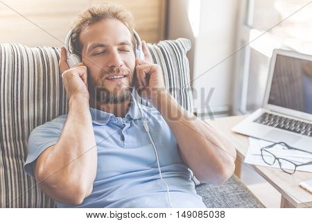 Handsome young businessman in casual clothes is listening to music in headphones while lying with closed eyes on couch at home
