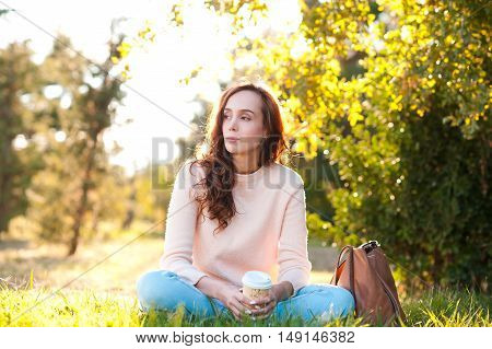 Beautiful giel 20-24 year old resting in park drinking coffee sitting on green grass outdoors. Looking away. Wearing casual clothes: knitted sweater and denim pants.