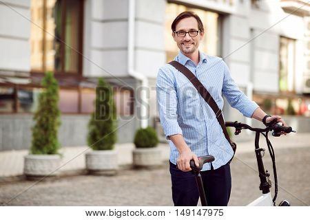 Happy ride. Handsome bespectacled man smiling and keeping bike while standing in the street.