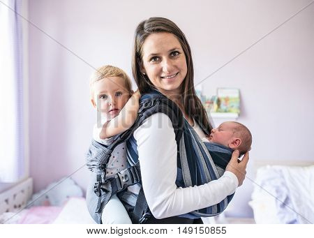 Beautiful young mother with her newborn son in sling and her daughter in baby carrier at home