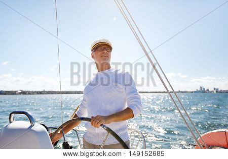 sailing, age, tourism, travel and people concept - happy senior man in captain hat on steering wheel and navigating sail boat or yacht floating in sea