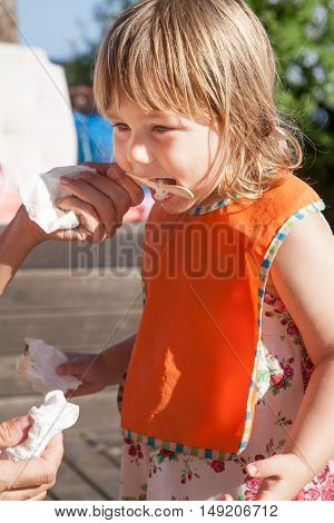 portrait of three years old blonde child with orange bib eating enjoying white ice cream from wooden stick in woman hand