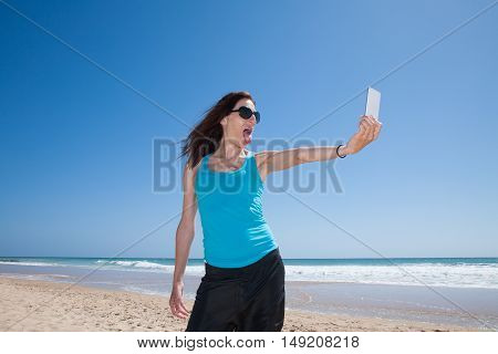 portrait of brunette woman with black sunglasses and blue cyan shirt teasing sticking out tongue making selfie with mobile phone smartphone at beach with sea behind