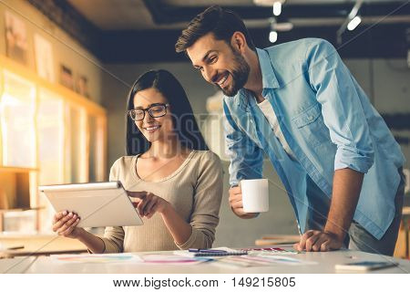 Beautiful couple of designers in casual clothes is using a digital tablet talking and smiling while working in studio