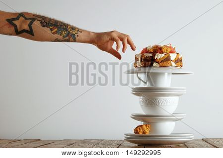 tattooed man's hand reaching out to grab a piece of cake balanced on a pyramid of white teaware