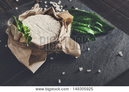 Cheese delikatessen closeup on black stone surface and wood. Camembert or brie circle in brown kraft paper decorated with basil, soft toning