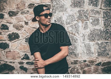 Always cool and trendy. Handsome young African man in cap  standing against the stoned wall and looking away