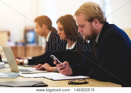 Young man is reading text message on mobile phone while is sitting in business school with his female partner what working on laptop computer. Male is searching information on portable cell telephone