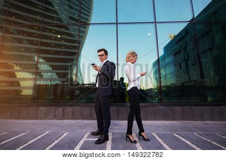 Young man and woman proud CEO are checking e-mail on smart phones while are standing outside company where they work. Two proud economist are reading financial news via cellphone during work break