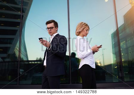 Man and woman successful business owner are using their mobile phones outdoors in urban setting. Two skilled managers are searching information on web page via phone before meeting with boss