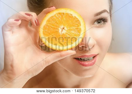 Young beautiful woman with perfect skin holding a slice of orange in front of her eyes and smiles looking ahead. Half-turn face. Gray background