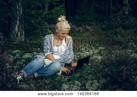 Young woman sitting on the grass in the forest and looking computer. Woman using laptop at outdoor.