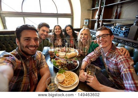 people, leisure, friendship and technology concept - happy friends taking selfie, drinking beer and eating snacks at bar or pub