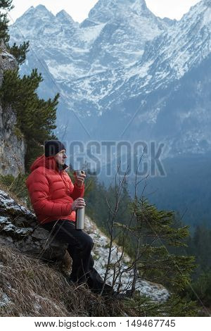 Man in winter mountains is resting and drinking from vacuum flask metal cup