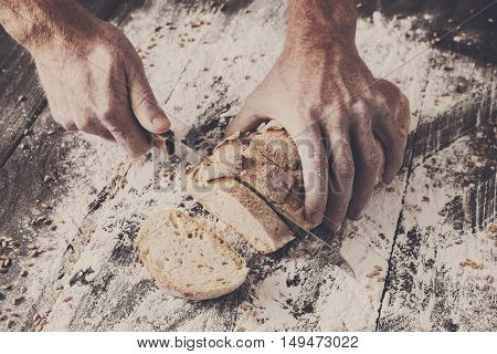 Baking and cooking concept background. Hands of baker cutting bread loaf with knife on rustic wooden table sprinkled with flour. Stained dirty hands of cook.