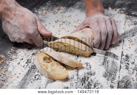 Baking and cooking concept background. Hands cutting bread loaf with knife on rustic wooden table sprinkled with flour. Stained dirty hands of baker.