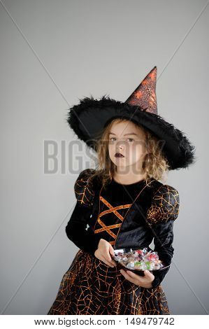 Girl in costume for Halloween. She in image the evil sorcerer. On the girl black-orange dress and a big hat. Girl holds a plate with candies and thoughtfully looks afar. Children adore Halloween.