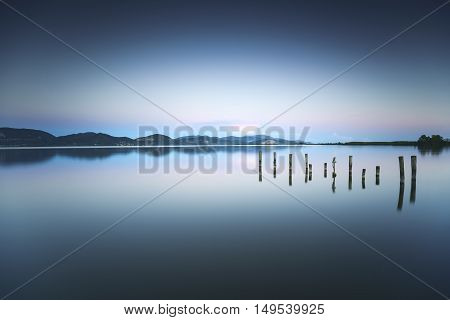 Wooden pier or jetty remains on blue lake sunset and sky reflection water. Long exposure Versilia Massaciuccoli Tuscany Italy.