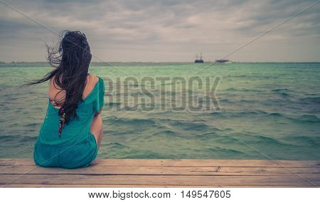 Closeup back view of long hair woman sitting in blue beach dress looking out towards blue ocean and sky. Retro style color tones.