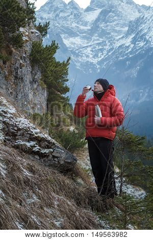 Mountaineer at winter snowy peaks background is resting and drinking from vacuum flask metal cup