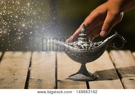 Beautiful antique metal lamp in true Aladin style, hand touching and animated star dust coming out, sitting on wooden surface.