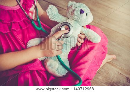 Child playing doctor or nurse with plush toy bear with bright sunlight at home. Happy girl listens a stethoscope to toy. Playful girl role playing. Vintage tone effect.