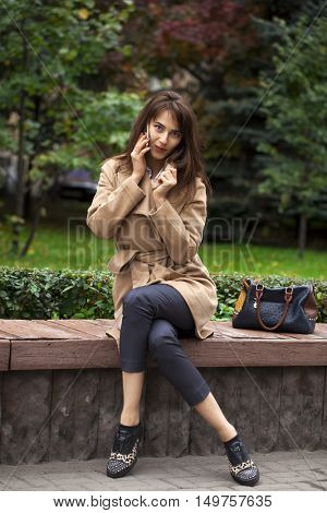 Young beautiful brunette woman in beige coat sitting on a bench in the autumn park