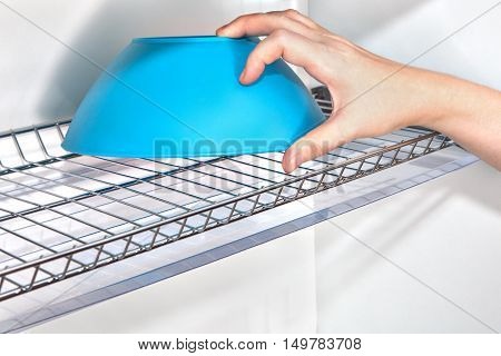 Close-up womans hand takes a blue bowl from the wall kitchen cupboard with wire plate drainer inside.