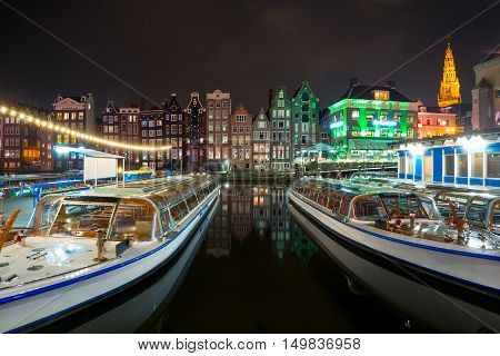 Beautiful typical Dutch dancing houses, Oude Kerk church and tourist boats at the Amsterdam canal Damrak at night, Holland, Netherlands.
