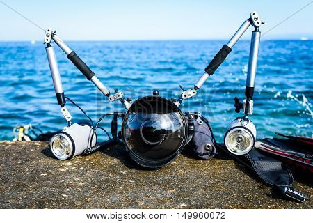 Professional underwater photography equipment for DSLR camera with strobes. Wide angle dome for fish-eye dual flashes with arms in aluminium casing on a pier with blue sea in the background.
