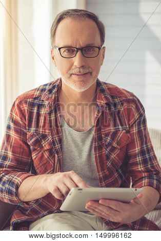 Handsome mature man in eyeglasses is using a digital tablet looking at camera and smiling while sitting on couch at home
