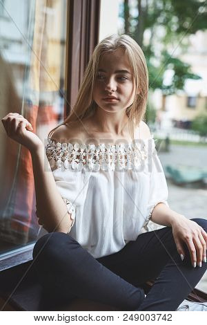 Young Blondie Girl In Trendy Clothes Sitting On The Window Sill In Coffeeshop.