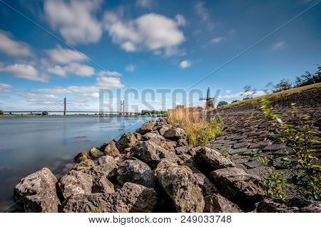 Dutch River Landscape With View On A Bridge, Traditional Thatched Windmill And A Dike With Basalt Bl