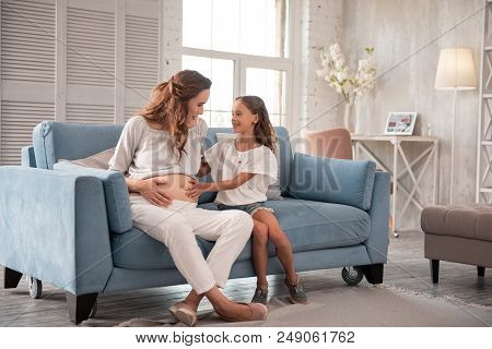 Comfortable Sofa. Cute Little Daughter Sitting On Comfortable Sofa Near Pregnant Mother